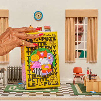 A hand holds Le Puzz’s The Flumpsteins 81 Piece Puzzle, featuring cartoon faces on a bright yellow box in a miniature room with tiny furniture and a checkerboard floor, as a smiling person and Snowball the cat peek through the window.