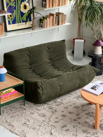 A cozy living room with a green Ligne Roset Togo 2 Seater loveseat, pleated fabric design, a light patterned rug, wooden coffee table with books, green side table, houseplants, and shelves displaying books and artwork.