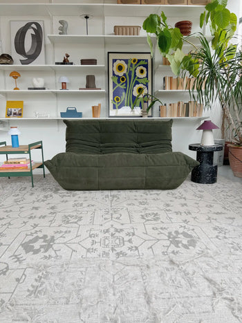 A modern living room with a Ligne Roset Togo, 2 Seater sofa by Ligne Roset featuring pleated upholstery, a patterned rug, potted plants, white shelving with books and decor, and vibrant floral wall art.