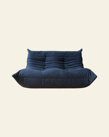 The Ligne Roset Togo, 2 Seater by Ligne Roset features modern, deep blue, tufted upholstery and a low, wide seat that channels mid-century style, shown armless against a plain beige background.