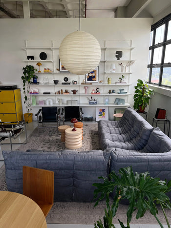 A modern living room featuring the Ligne Roset Togo 2 Seater by Ligne Roset in gray, a round white pendant lamp, open wall shelves with decor, plants, a yellow cabinet, and large windows for natural light—all with mid-century flair.
