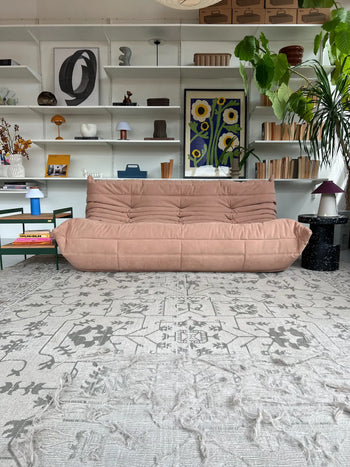A modern living room with a Ligne Roset Togo, 3 Seater sofa in pink, patterned gray rug, white book-filled shelves, abstract art, plants, and colorful table lamps—all blending mid-century accents.