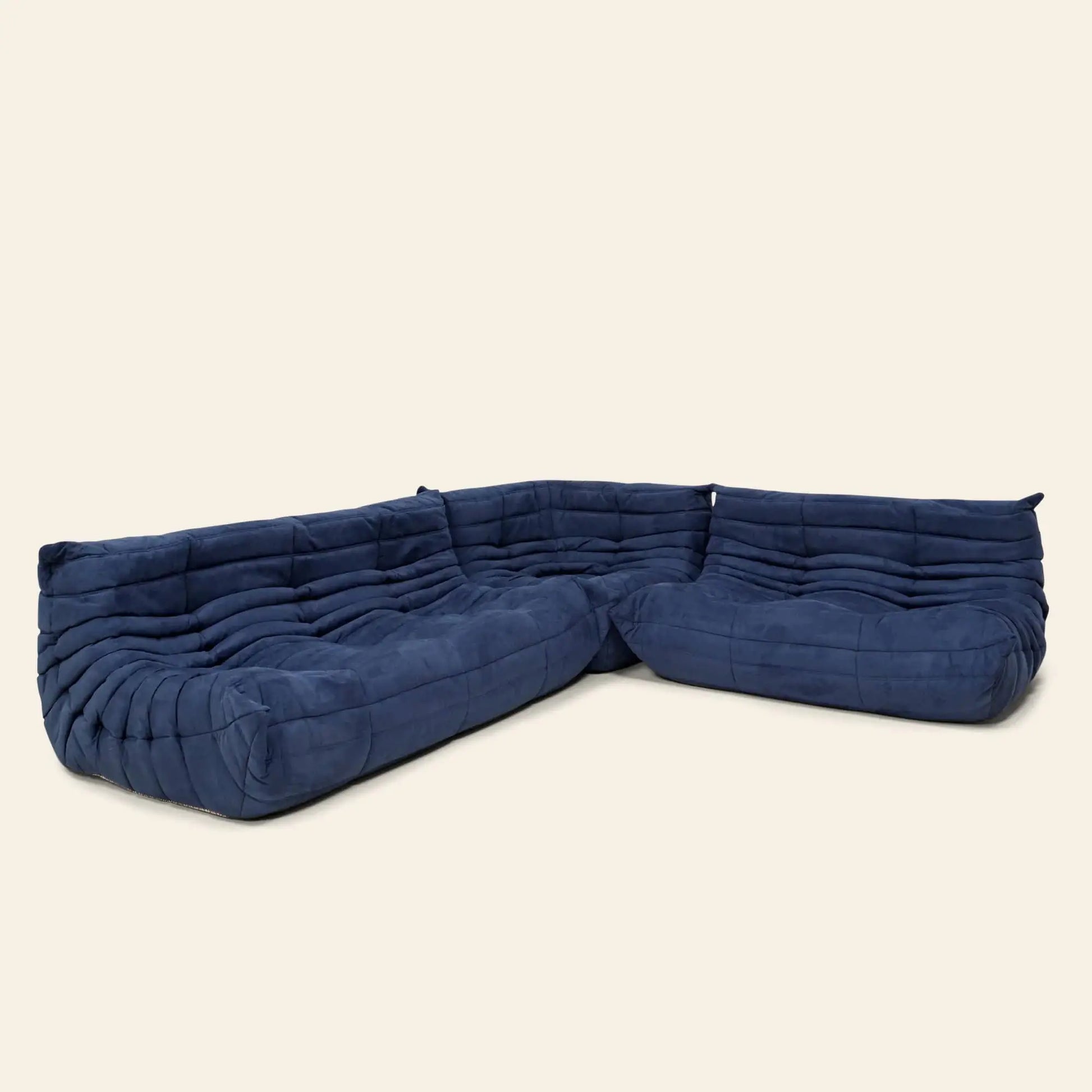The Ligne Roset Togo, 3 Seater by Ligne Roset is a dark blue, low-profile L-shaped sectional with plush ruched upholstery and modular design inspired by the iconic Togo sofa, featuring a modern minimalist look against a light backdrop.