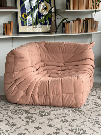 A plush tan Ligne Roset Togo Corner Seat with a quilted, ruched design sits on a patterned rug before white shelves filled with books and modern art.