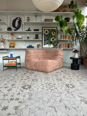 A cozy living room features a tan Ligne Roset Togo Corner Seat, mid-century style, on a patterned gray rug. A green side table stands beside bookshelves with decor, books, and plants. A floral art print and large potted plant sit in the background.