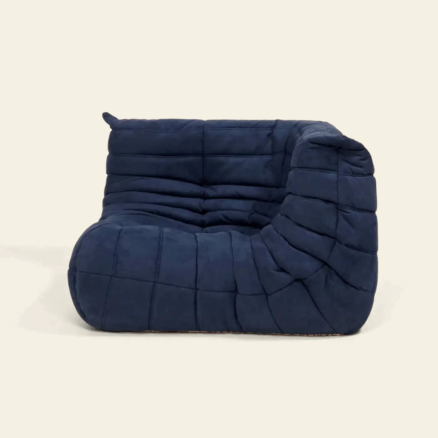 The Ligne Roset Togo Corner Seat by Ligne Roset is an armless, navy blue lounge chair with pleated fabric, soft rounded edges, and mid-century-inspired quilted cushioning, set against a plain beige background.
