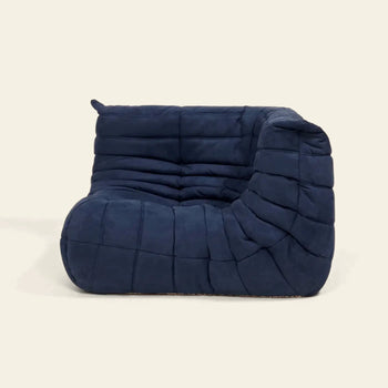 The Ligne Roset Togo Corner Seat by Ligne Roset is an armless, navy blue lounge chair with pleated fabric, soft rounded edges, and mid-century-inspired quilted cushioning, set against a plain beige background.