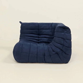 The Ligne Roset Togo Corner Seat by Ligne Roset is a modern, armless lounge chair with dark blue pleated upholstery, a quilted textured look, low seat, and sculpted curved backrest inspired by mid-century design on a light background.