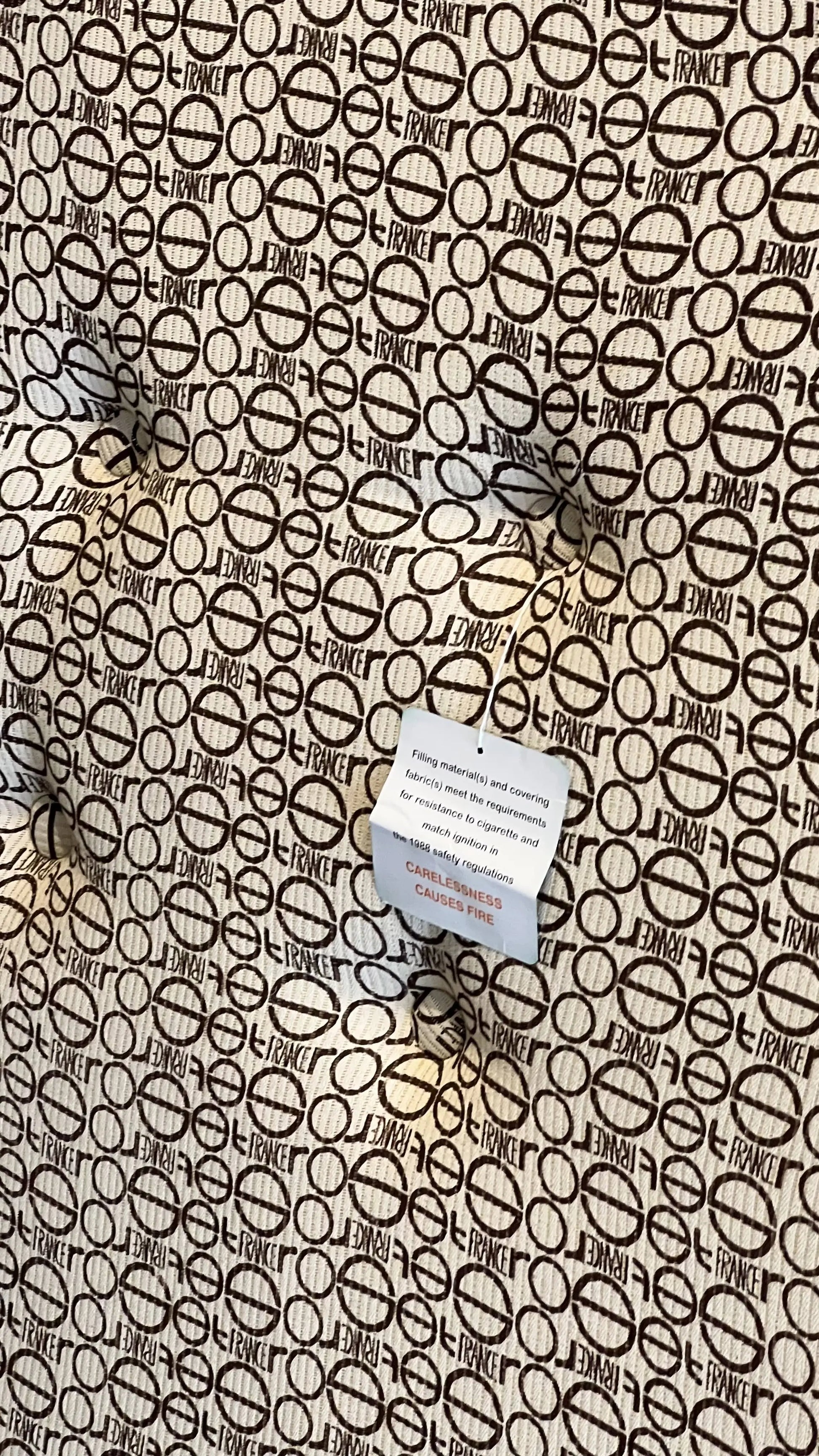 A close-up of the Ligne Roset Togo, Corner Seat by Ligne Roset shows brown fabric with repeating LE FRANCE text and crossed-out circles. A white care tag is attached, highlighting its mid-century modular-inspired design.