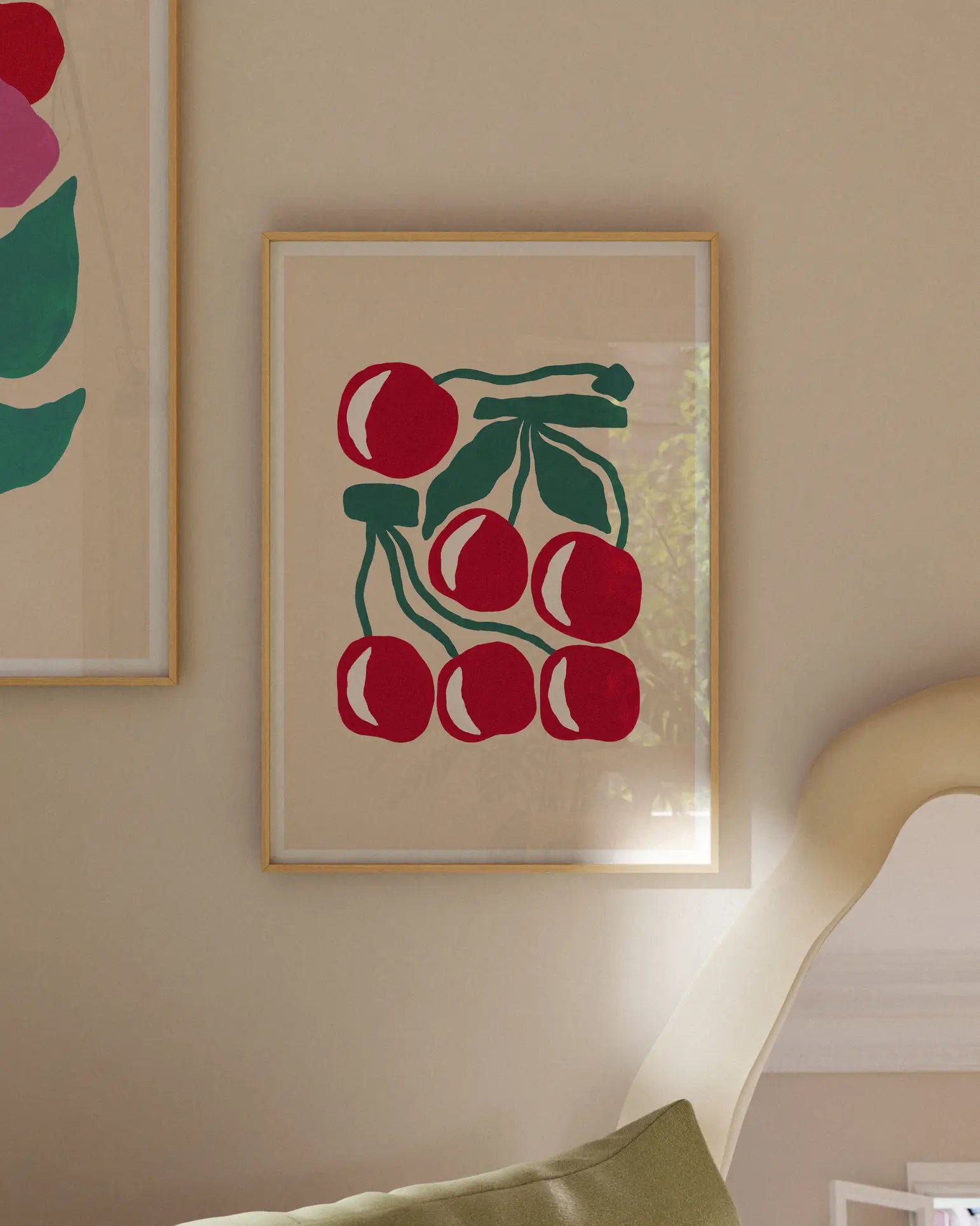 Liv Lee Cherries (Cream) by Liv Lee is a minimalist A3 giclée print featuring six red cherries with green leaves on archival Hahnemühle paper, framed and displayed on a beige wall beside a chair and another vibrant artwork.