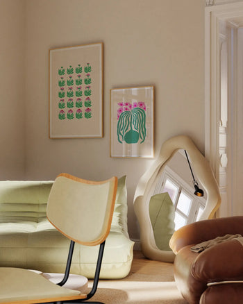A cozy living room corner features a green sofa, modern cream chair, wavy floor mirror, and two Liv Lee Daisy Frames art prints by Liv Lee on Hahnemühle paper adorning the beige wall in soft natural light.
