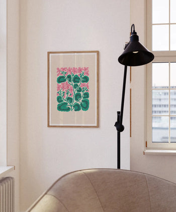 The Liv Lee Geraniums print by Liv Lee, featuring abstract pink geraniums and green leaves, hangs on a white wall near a window with a black floor lamp and part of a beige armchair in view.