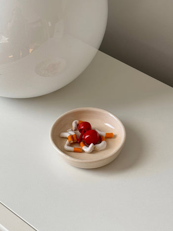 A small beige MIRYO Miss Cherries and Cigs Ashtray holds colorful, toy-like mushroom accents, adding a unique decorative touch to a white surface near a round, reflective lamp.