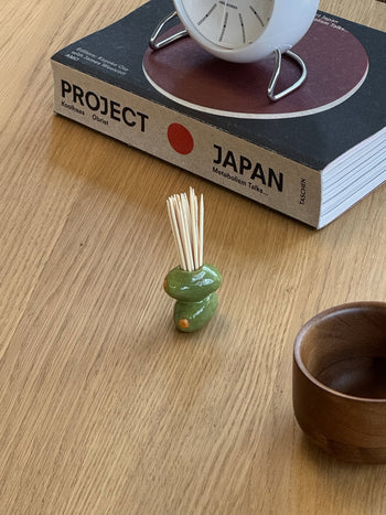 The MIRYO Olive Toothpick Holder by MIRYO, filled with toothpicks, a wooden bowl, and the large book Project Japan sit on a wooden table. A white clock rests atop the book.