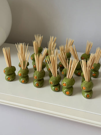 Several MIRYO Olive Toothpick Holders, handcrafted to resemble green olives with pimentos and designed to hold toothpicks upright, are arranged on a white tray against a neutral background.