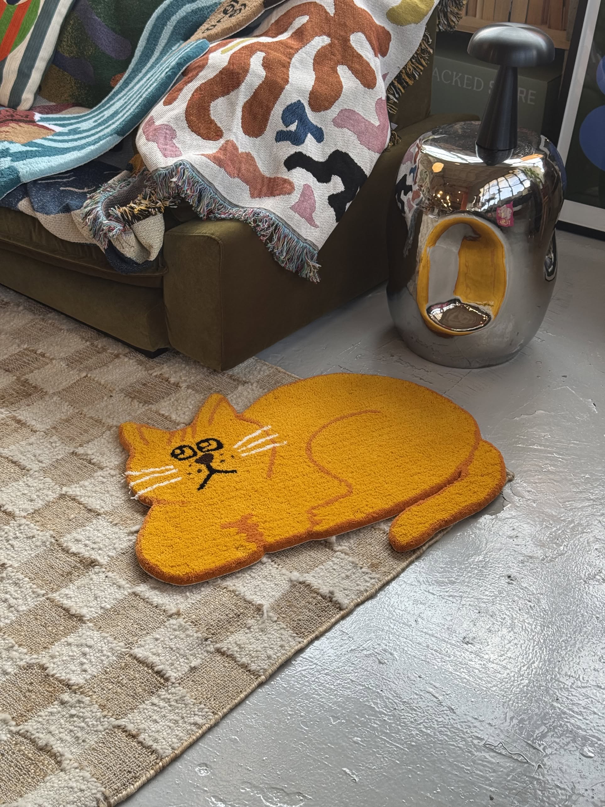 The MIYOSHI RUG Shiomi Wada x MIYOSHI RUG - Cat Rug A in orange rests on a checkered carpet near a green couch with a colorful throw and a shiny silver cat-shaped side table featuring a yellow interior.