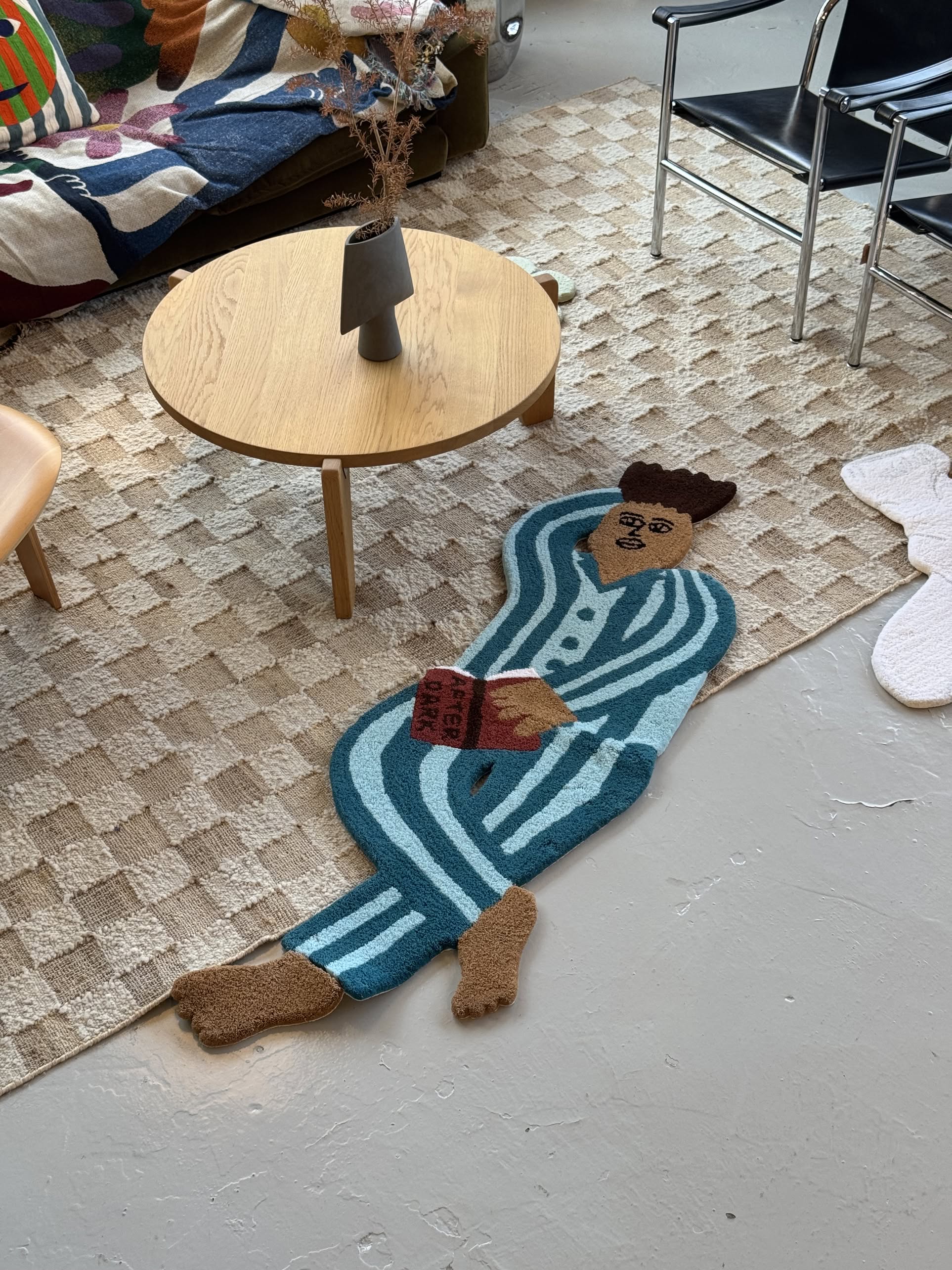The MIYOSHI RUG Shiomi Wada x MIYOSHI RUG - Your Friend Rug features a blue-and-white pajama-clad human figure holding a red book, displayed on a beige carpet near modern furniture in a stylish living room.