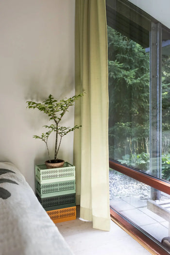 A small potted plant rests on Made Crate Midi eco-friendly folding crates from Made Crate, crafted from recycled plastic, by a floor-to-ceiling window with light green curtains overlooking a lush garden. Part of a couch appears on the left.