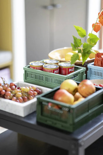 A table displays Made Crate Mini folding crates by Made Crate, filled with canned tomatoes, apples, a marble bowl of grapes, and a vase with leafy branches; extra produce is stacked conveniently in the background.