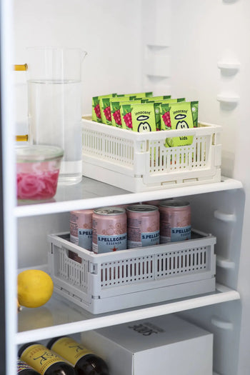 A neatly organized fridge shelf features boxed juices in Made Crate Mini eco-friendly folding crates by Made Crate, San Pellegrino cans in a gray recycled plastic crate, a glass jug of water, pickled onions in a bowl, a lemon, and conveniently stacked bottles.