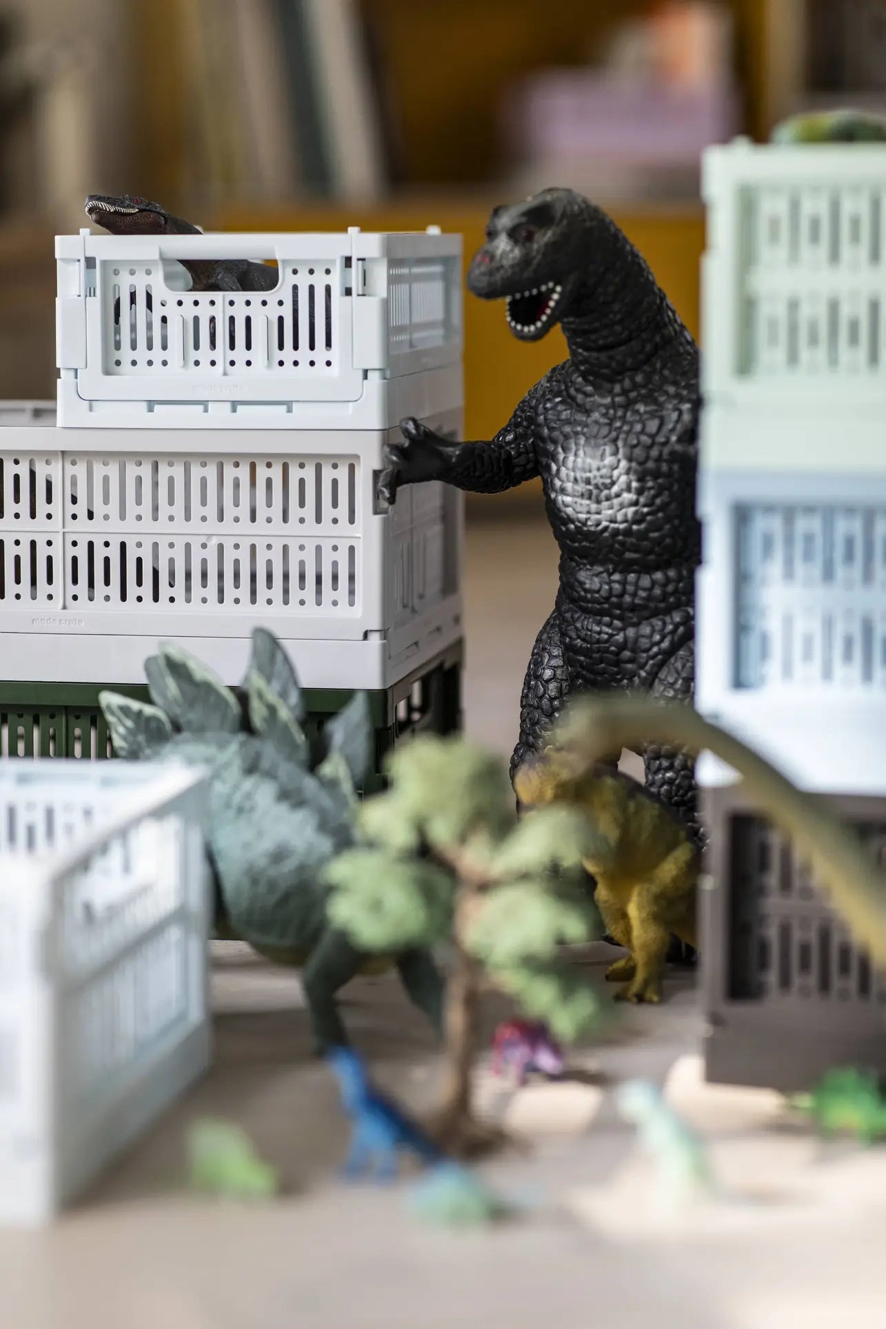 Toy dinosaurs, including a large Godzilla and a stegosaurus, roam among mini buildings and trees atop Made Crate Mini eco-friendly folding crates by Made Crate, made from recycled plastic for stacking convenience in this playful tabletop city.