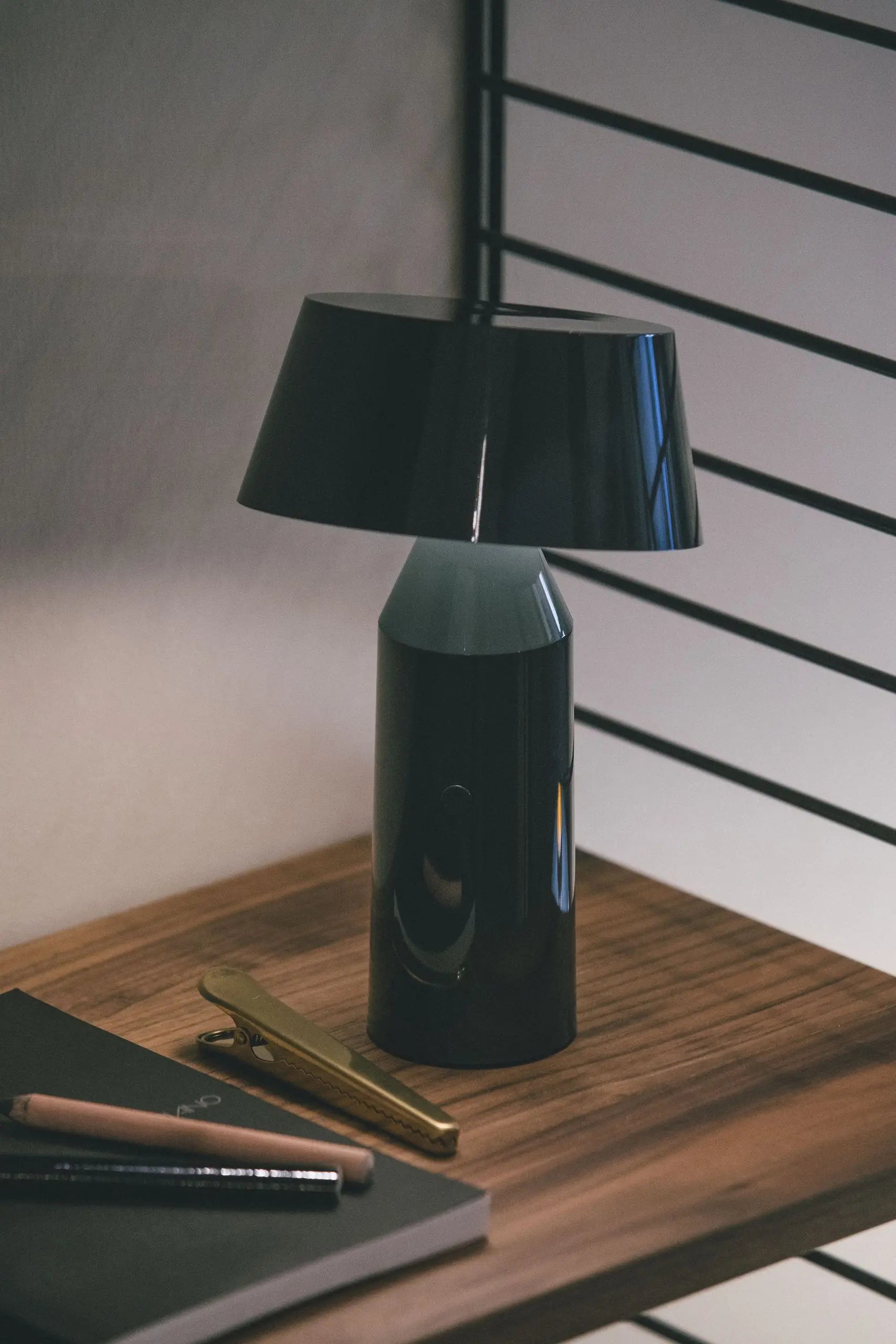 The Marset Bicoca Portable Table Lamp by Marset sits on a wooden surface beside a closed notebook, pencil, and gold clip, casting soft light and gentle shadows while black metal bars accent the background.