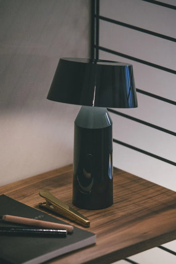The Marset Bicoca Portable Table Lamp by Marset sits on a wooden surface beside a closed notebook, pencil, and gold clip, casting soft light and gentle shadows while black metal bars accent the background.