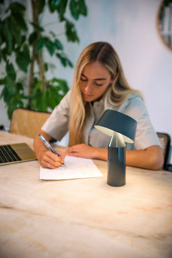 A woman with long blonde hair writes at a desk with a laptop, pen, and the Marset Bicoca Portable Table Lamp from Marset, adding charm to her workspace as green plants decorate the background.