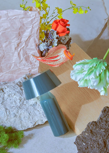 A Marset Bicoca Portable Table Lamp by Marset sits stylishly on its side amid textured rocks, pastel pink paper, a wooden board, and colorful flowers like a red rose and green tulip for an artistic, eclectic look.