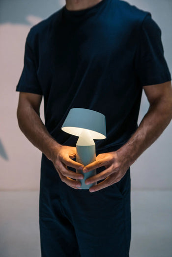 A person in a dark outfit holds the Marset Bicoca Portable Table Lamp, casting a soft, warm glow. The minimal, blurred background accentuates the modern design of this portable lamp by Marset.