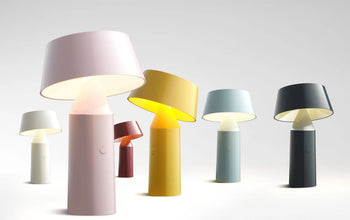 A row of Marset Bicoca Portable Table Lamps by Marset in matte pink, red, yellow, blue, and black are displayed on a light surface. These stylish lamps cast a warm glow, making them perfect for any space.