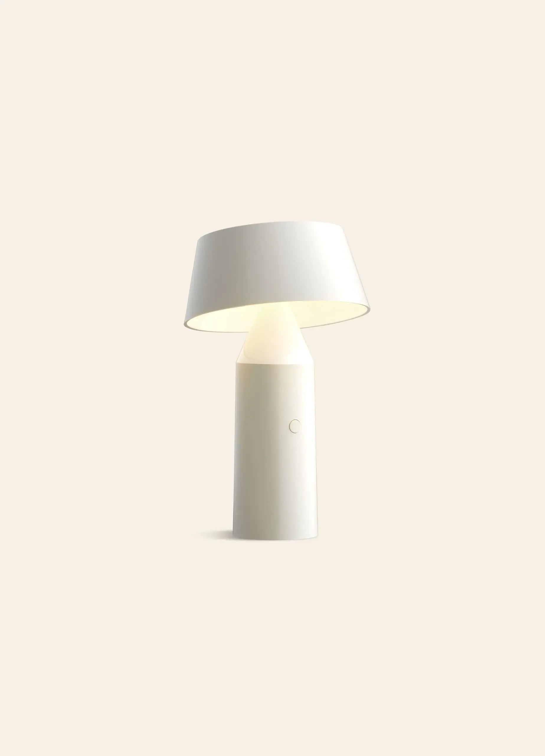 The Marset Bicoca Portable Table Lamp by Marset features a modern, minimalistic design with a cylindrical base and slightly angled wide lampshade, set against a plain light beige background.