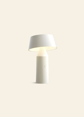 The Marset Bicoca Portable Table Lamp by Marset features a modern, minimalistic design with a cylindrical base and slightly angled wide lampshade, set against a plain light beige background.