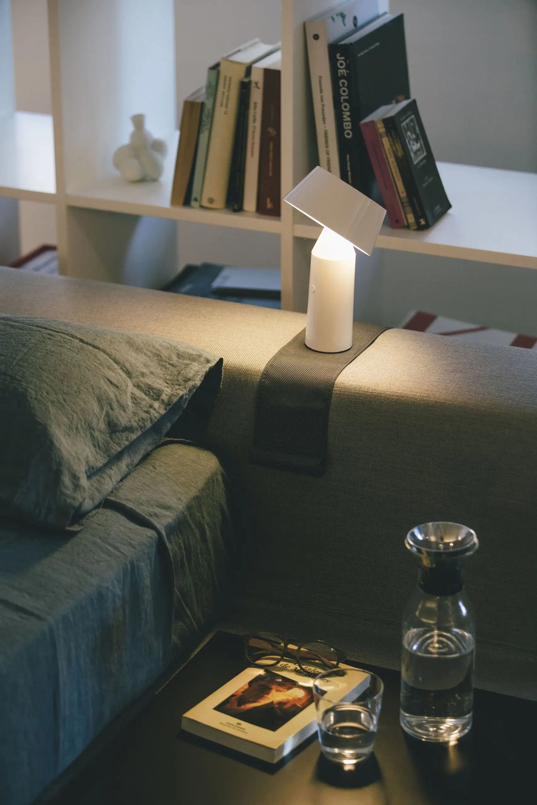 A modern bedside scene features the Marset Bicoca Portable Table Lamp by Marset on a gray headboard, books on a white shelf, a glass carafe with water and glass, eyeglasses, and a photo-covered book on a black nightstand.