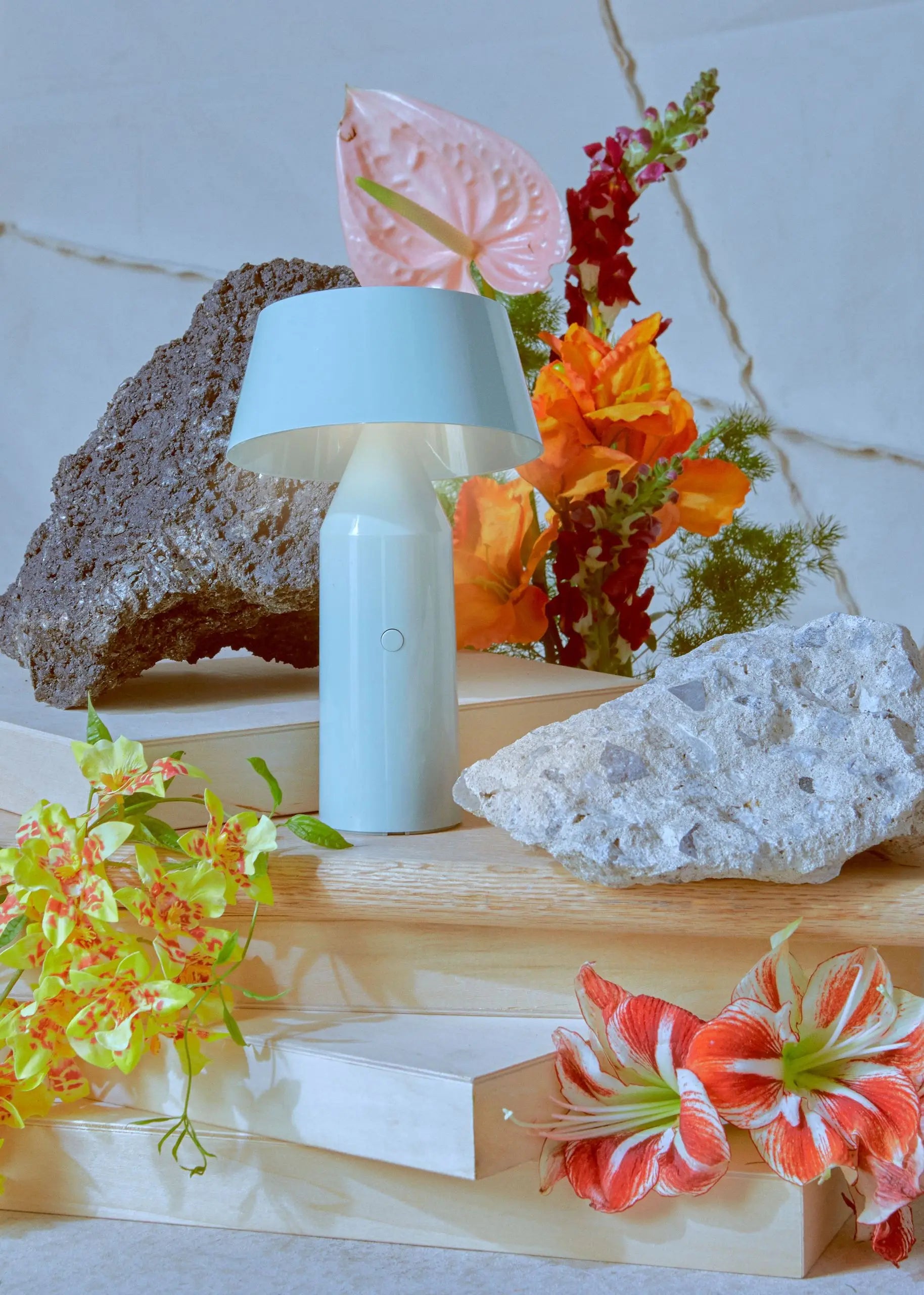 The Marset Bicoca Portable Table Lamp by Marset in light blue is showcased on wooden platforms, surrounded by vibrant flowers and two textured rocks, set against a light marble background with visible veining.