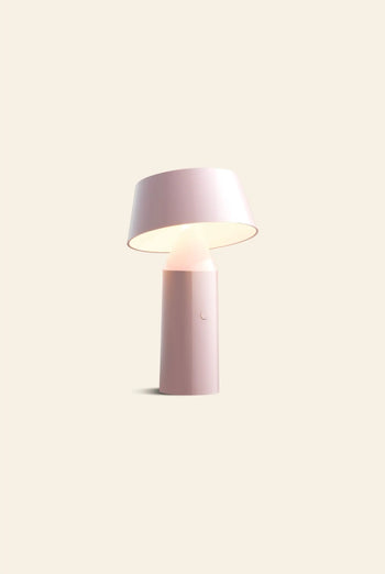 The Marset Bicoca Portable Table Lamp by Marset, featuring a modern minimalist design with a cylindrical base and a tilted wide lampshade, softly illuminates against a plain beige backdrop, bringing stylish flair to any space.