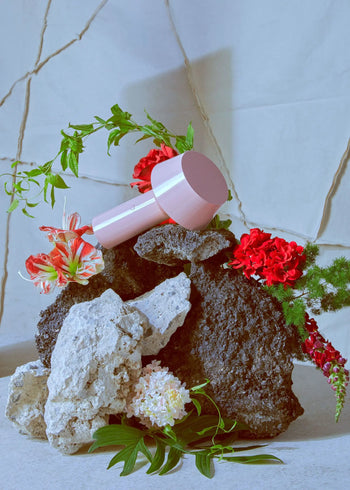 A pink hairdryer rests on rocks amid vibrant flowers and green leaves, with a Marset Bicoca Portable Table Lamp by Marset, all set against a light, textured background featuring visible cracks.