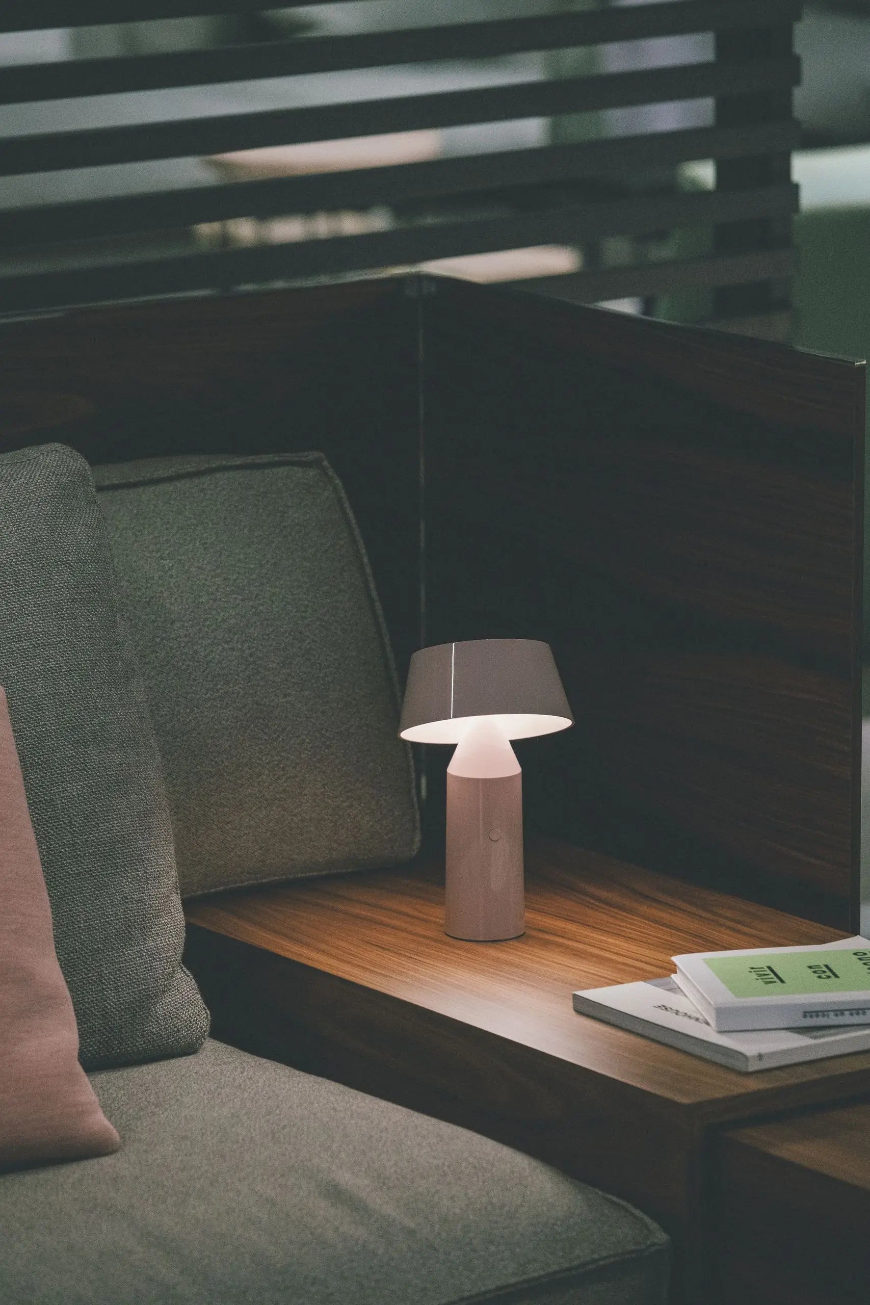 The Marset Bicoca Portable Table Lamp by Marset casts a soft glow on a wooden side table beside a gray sofa with a pink cushion, with a stack of books nearby and dark wooden panels in the background.