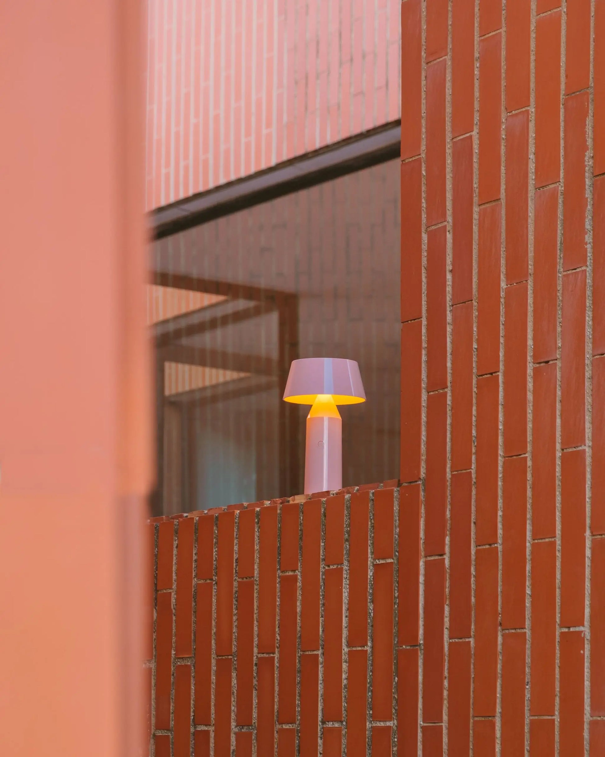 The Marset Bicoca Portable Table Lamp adds a warm, stylish glow to a ledge framed by red brick walls and large glass windows—an ideal accent piece from Marset for contemporary interiors.