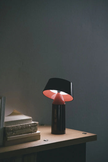 The Marset Bicoca Portable Table Lamp by Marset, in black and red, rests on a wooden surface near a stack of books, softly lighting up the dimly lit room.