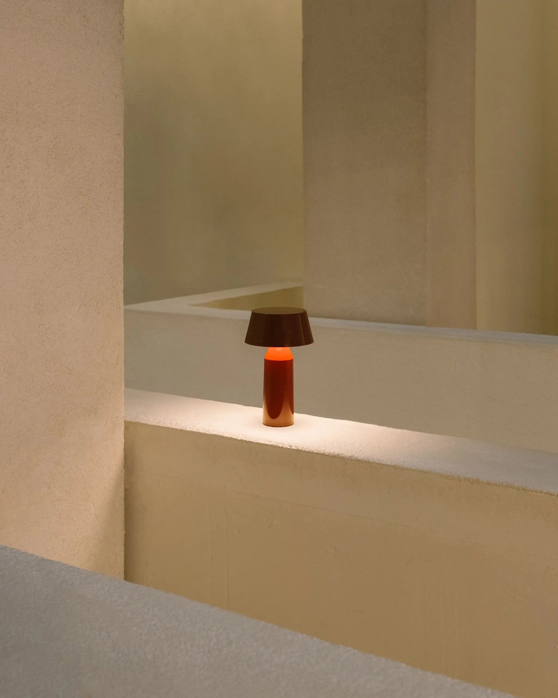 The Marset Bicoca Portable Table Lamp, featuring a brown shade and orange base, sits on a beige ledge amid minimalist cream walls and geometric architectural shapes.