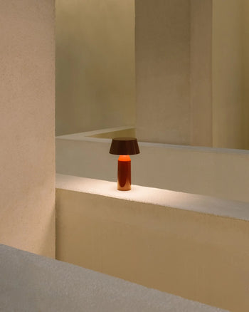 The Marset Bicoca Portable Table Lamp, featuring a brown shade and orange base, sits on a beige ledge amid minimalist cream walls and geometric architectural shapes.