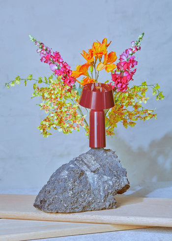 A vibrant floral arrangement with orange, pink, and yellow blooms emerges from a Marset Bicoca Portable Table Lamp by Marset atop rough, dark volcanic rock against a light background.