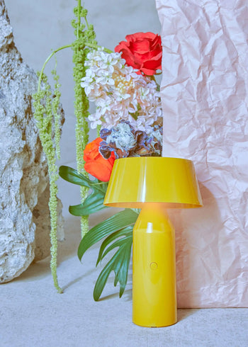 The Marset Bicoca Portable Table Lamp by Marset in vibrant yellow is displayed before a pink crumpled paper backdrop, accompanied by a stone and a colorful arrangement of red, purple, and white flowers with green leaves.