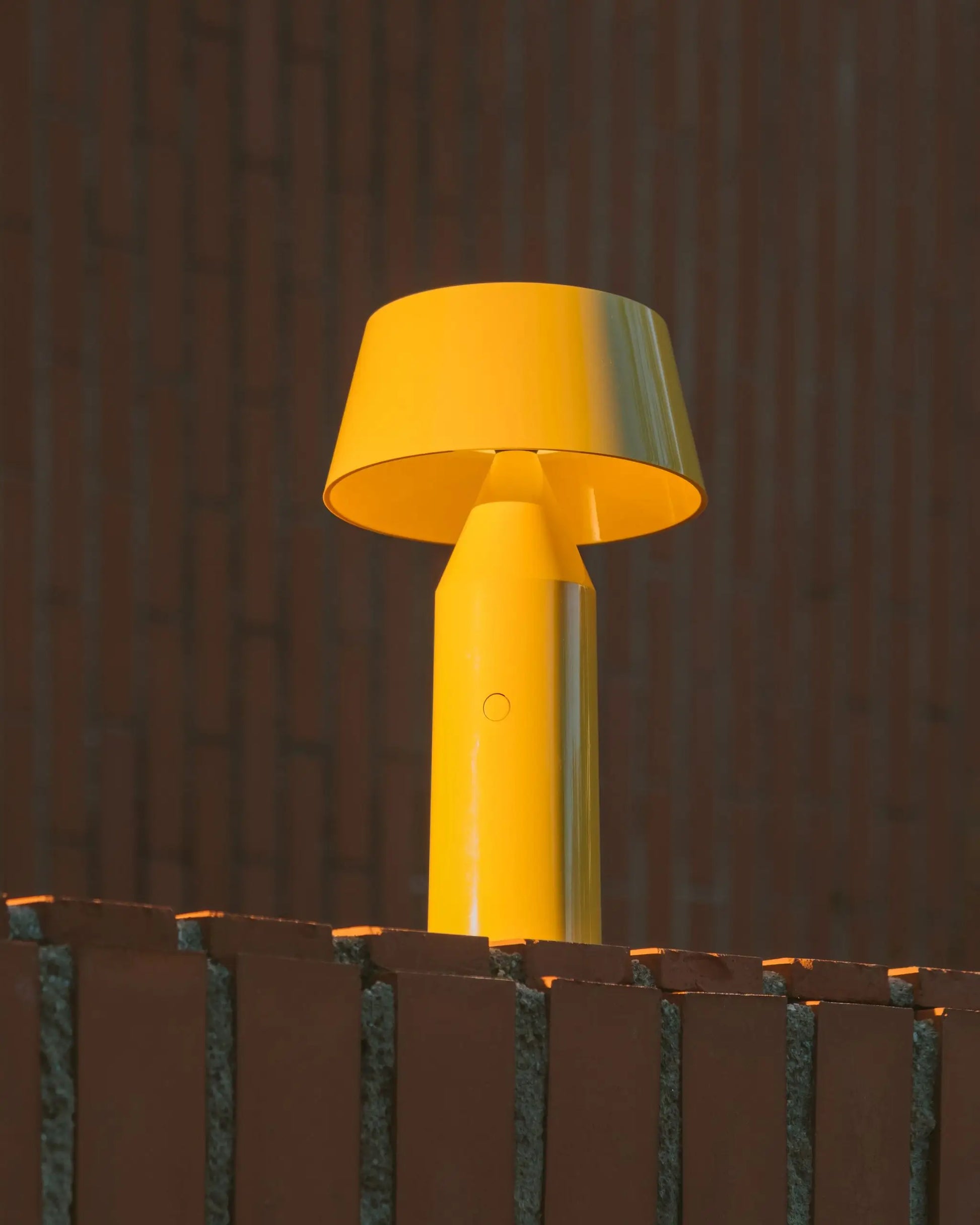 A vibrant yellow Marset Bicoca Portable Table Lamp with a wide shade rests on a red brick wall, set against a blurred red brick background.