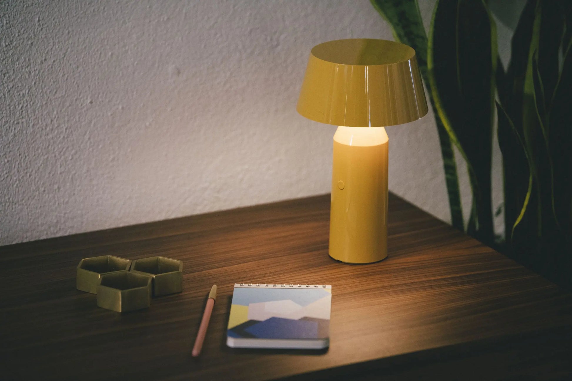 The Marset Bicoca Portable Table Lamp by Marset sits on a wooden surface beside a pink pencil, a geometric notebook, two hexagonal dishes, and a green plant in the background.