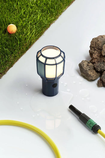 The Marset Chispa Portable Table Lamp by Marset, featuring a cage-like cover, stands on a wet white surface near a yellow garden hose—ideal for portable, rechargeable indoor or outdoor lighting.