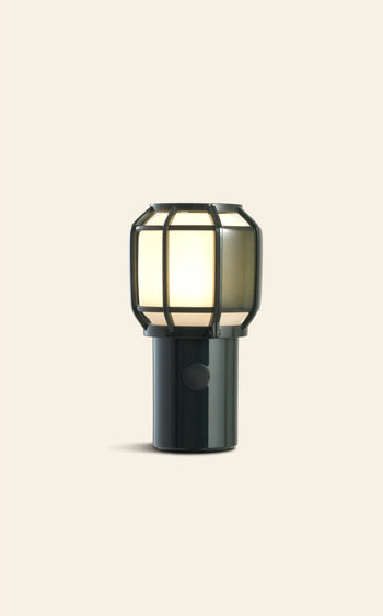 The Marset Chispa Portable Table Lamp by Marset features a chargeable black cylindrical base and a round frosted glass shade encased in a black metal cage, offering soft indoor-outdoor lighting against a plain beige background.