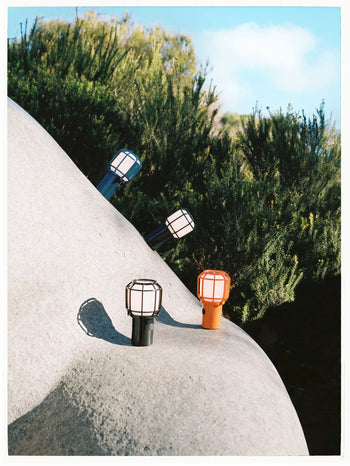 Four Marset Chispa Portable Table Lamps in black, white, blue, and orange sit outdoors on a rock among greenery under a partly cloudy sky, showcasing Marset’s stylish, chargeable design for indoor or outdoor portable lighting.