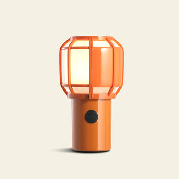 The Marset Chispa Portable Table Lamp by Marset features a modern orange design with a cylindrical base and cage-like cover, providing portable and rechargeable indoor-outdoor lighting against a simple light background.